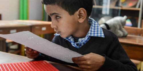 A visually disabled young student in a classroom holding a Braille sheet in his hands.